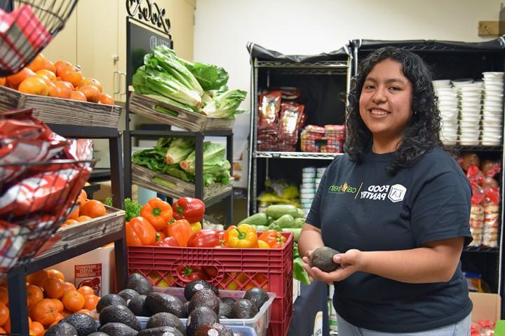A person wearing a Cal State LA Food Pantry t-shirt smiling while standing in front of bins of fresh produce.