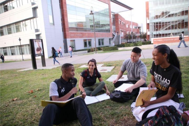 两个男学生和两个女学生坐在草坪上. 他们后面是红砖建筑